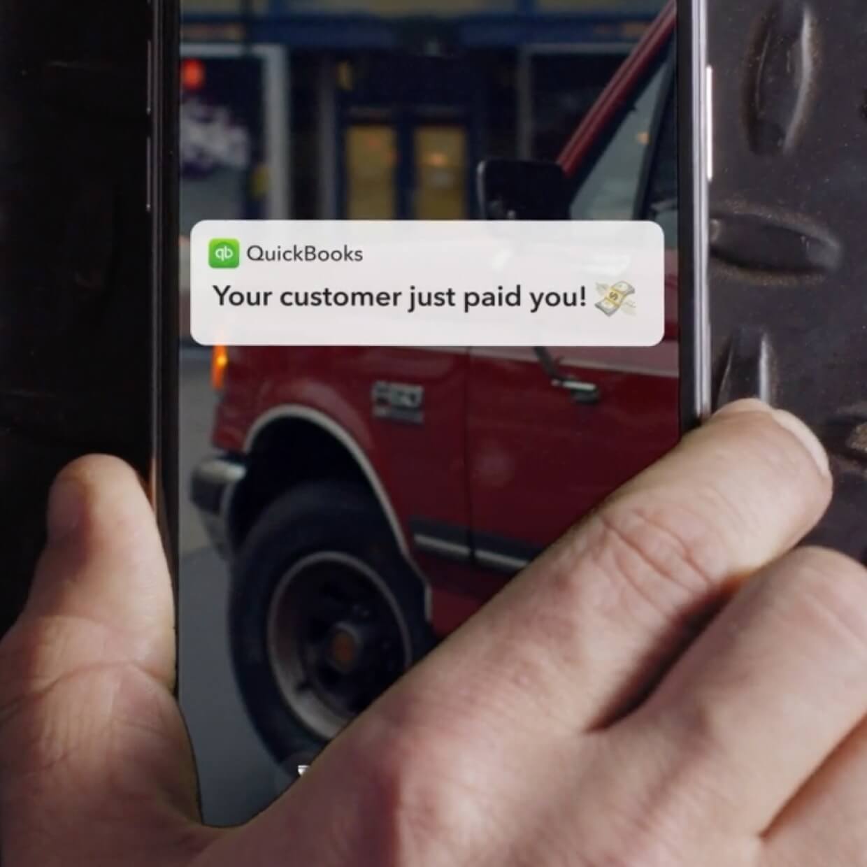 Hand holding a phone with a notification from QuickBooks that reads Your customer just paid you.